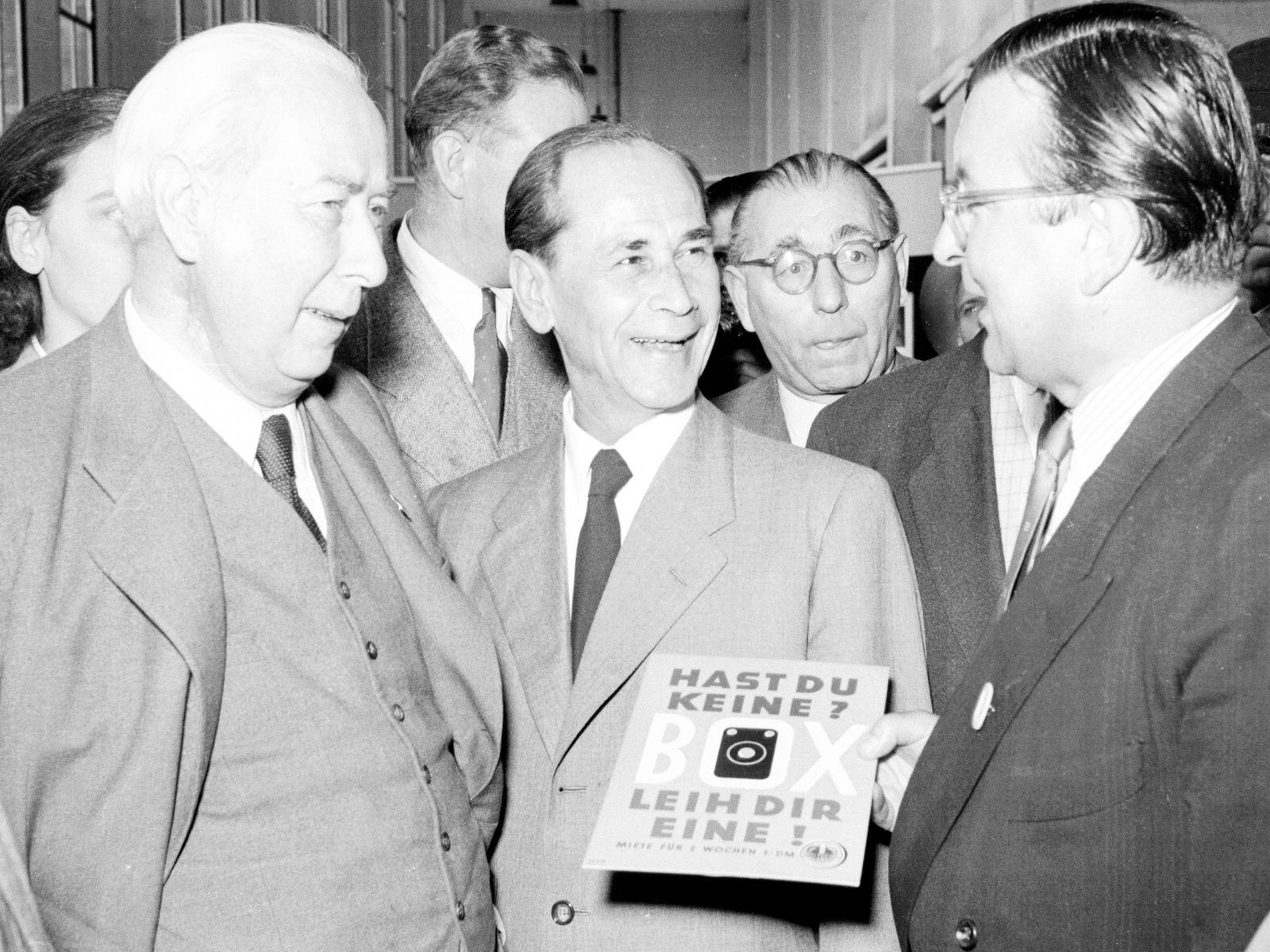 Bundes­präsident Theodor Heuss auf der Photo­kina 1952. Ver­treter der Foto­industrie zeigen Werbe­material zu der Werbe­kampagne Hast Du keine? -- Box -- Leih' Dir eine! (Foto: Dohm; StadtA Rt. S 105/5 Nr. 2123/30)