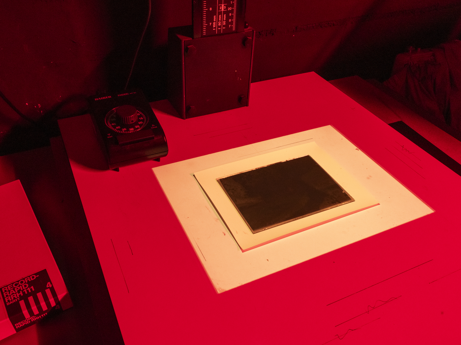 Farbbild (born-digital): Kopieren eines Glasplattennegativs im Kontakt auf Baryt-Fotopapier unter einem Vergrößerungsgerät im Fotolabor bei Rotlicht. Daneben liegt eine Schachtel Record-Rapid-Fotopapier von Agfa. (Foto: © Martin Frech, 12/2025)