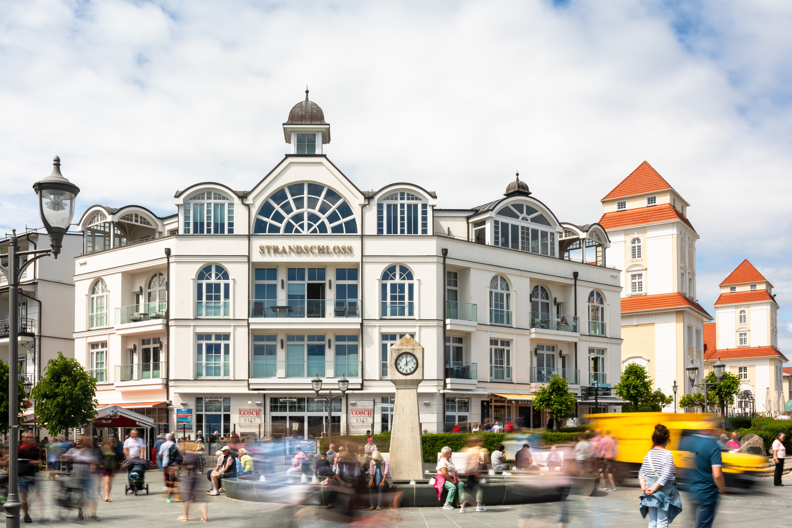 Standuhr mit Brunnen, Hauptstraße, Binz; i.H:.: Fassade Haus Strandschloss und Kurhaus; Passanten sind wegen der langen Belichtungszeit von 0,8 Sekunden teilweise verschwommen (Aufnahme mit Graufilter, ND 1.8; Foto: Martin Frech, 5/2024)